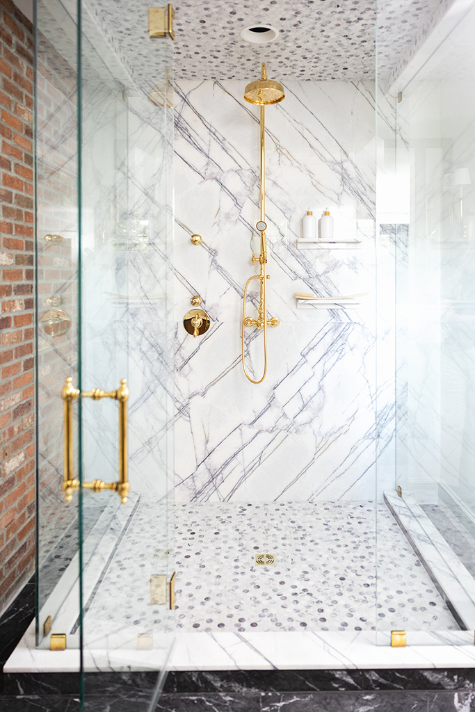 Detail of the unlacquered brass rain shower head and handheld sprayer against marble tile.